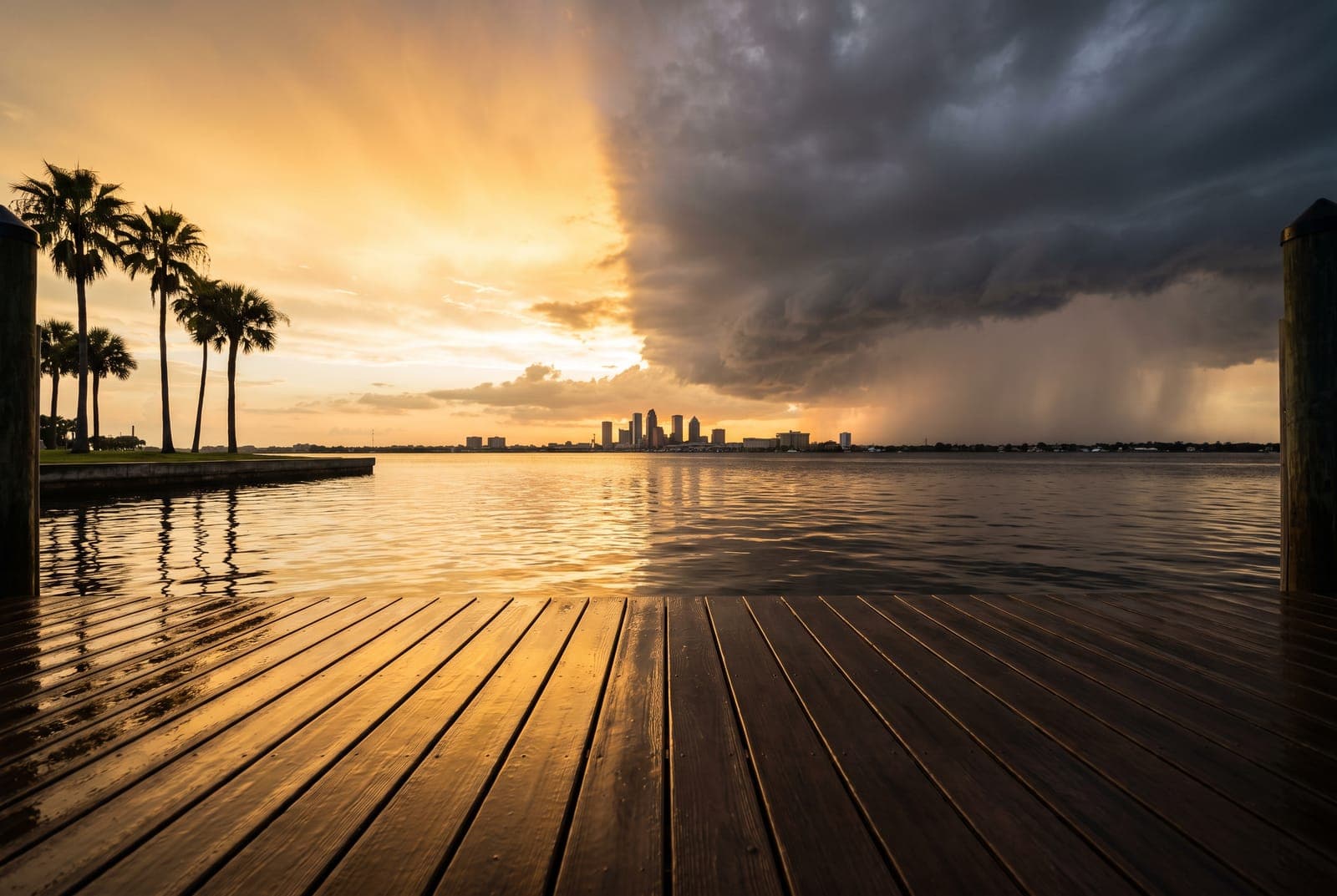 A storm front splitting the sky over Tampa Bay at sunset — golden light and silhouetted palms on the left, dark clouds and visible rain shafts falling over the downtown Tampa skyline on the right, seen from a rain-slicked wooden dock — El Niño winter 2026-27 Q4 and Q1 vacation rental forecast, Emperor Rentals